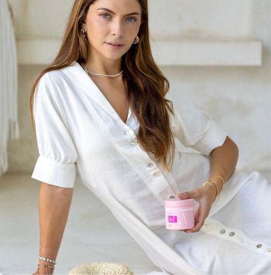 Woman in a white outfit holding a pink jar OF aZURE tAN fIRM AND TONE TANNING BUTTER in a neutral setting