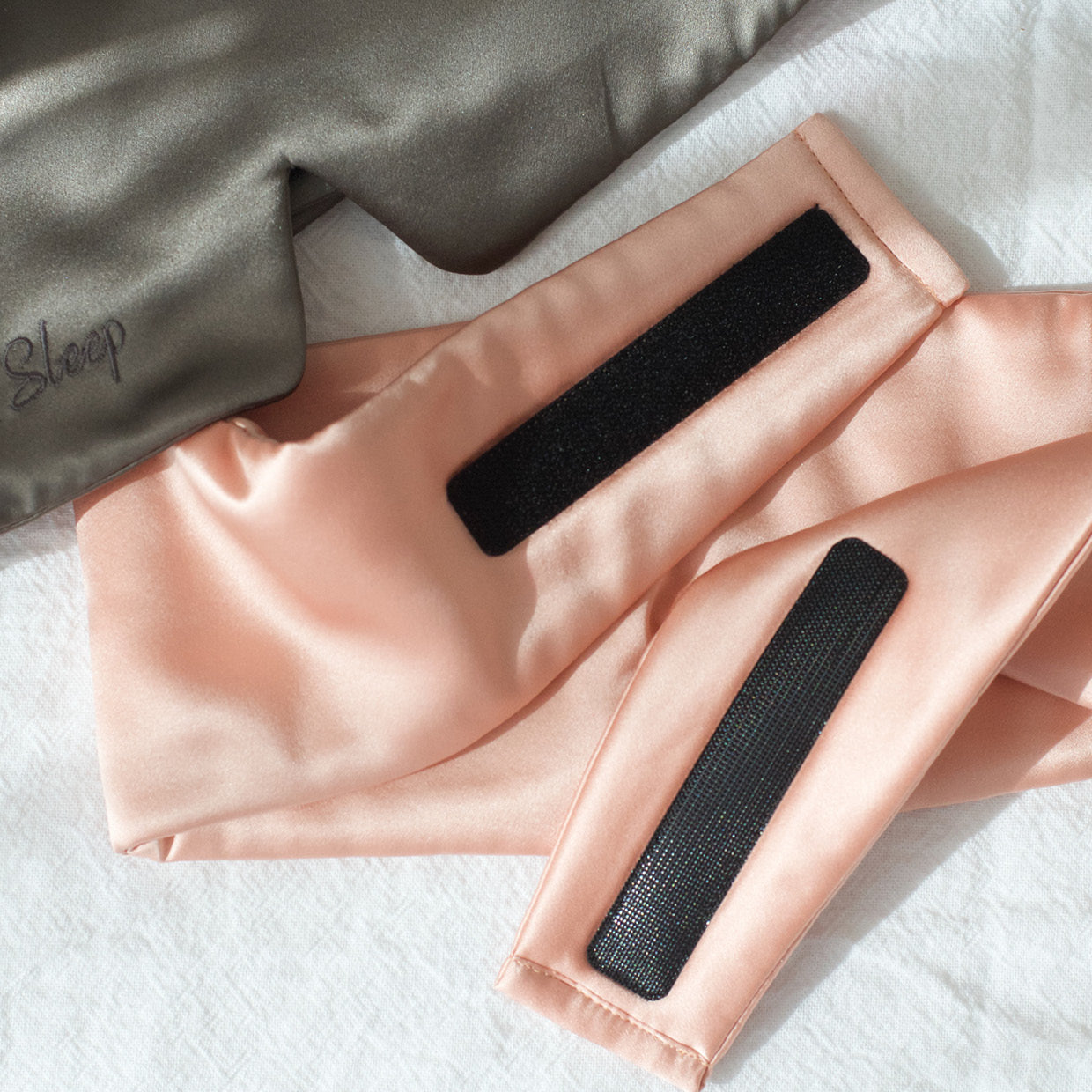 Charcoal Slate eye mask and blush pink eye mask on a white linen sheet. Slate eyemask is the font with the "Sleep" part of the embridored logo showing. Blush pink mask is the back showing the luxurious strong velcro that holds the mask together.