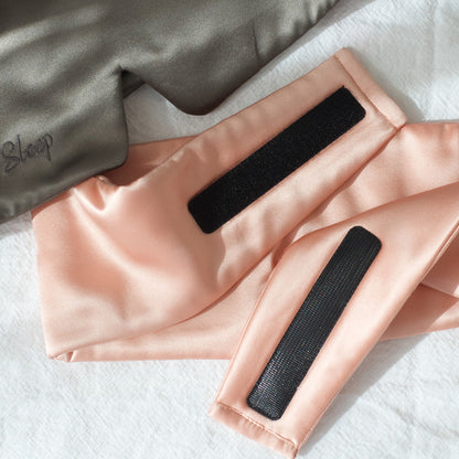 Charcoal Slate eye mask and blush pink eye mask on a white linen sheet. Slate eyemask is the font with the "Sleep" part of the embridored logo showing. Blush pink mask is the back showing the luxurious strong velcro that holds the mask together.
