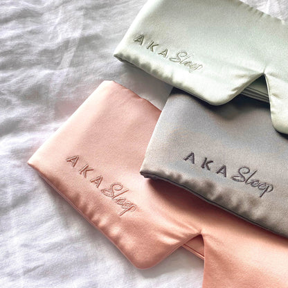 AKA Sleep Deep Sleep Mulberry Silk Eye mask in blush pink, sage green and slate grey lying on a white linen sheet