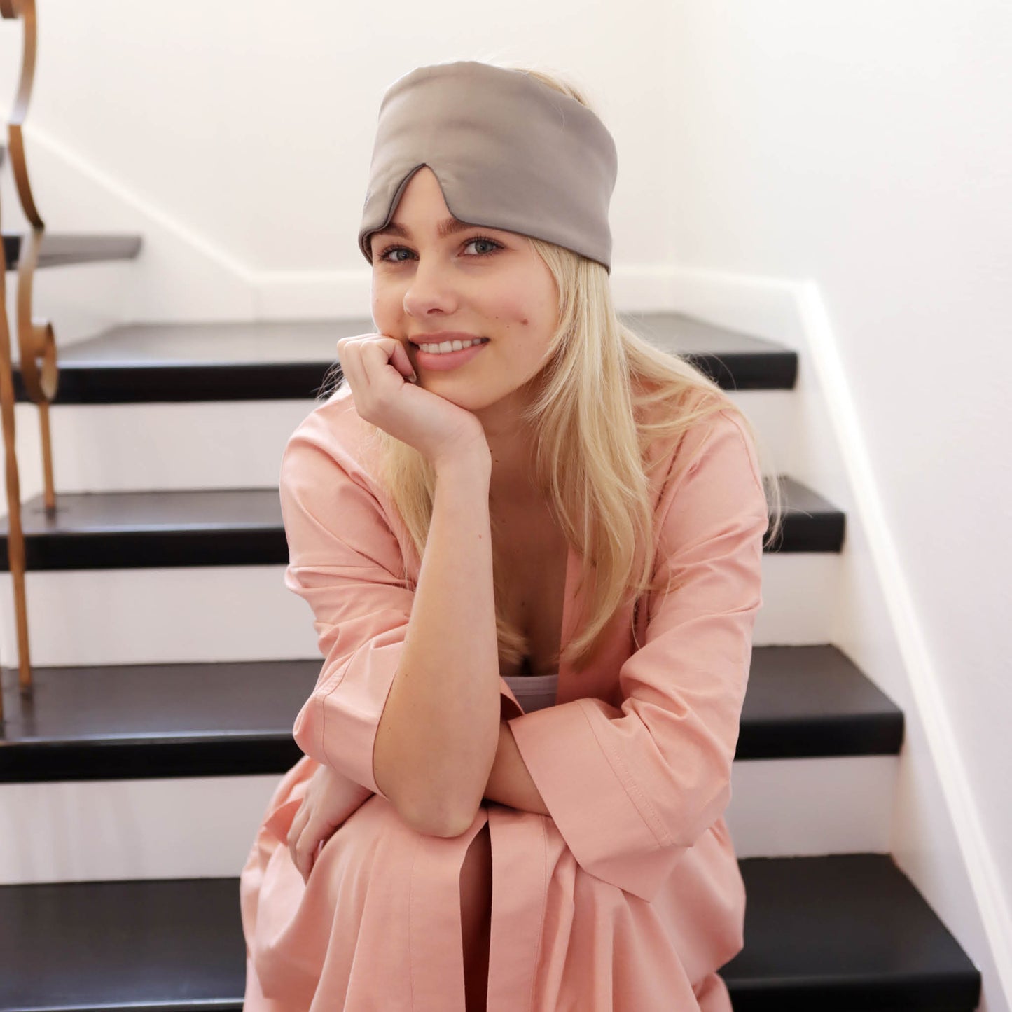 Beautiful blonde young lady is sitting on a curved white and dark wood staircase. She is wearing a peach linen robe and has the slate charcoal mask on the top of her head. Her head is gently resting on her hand as she looks straight at the camera. 