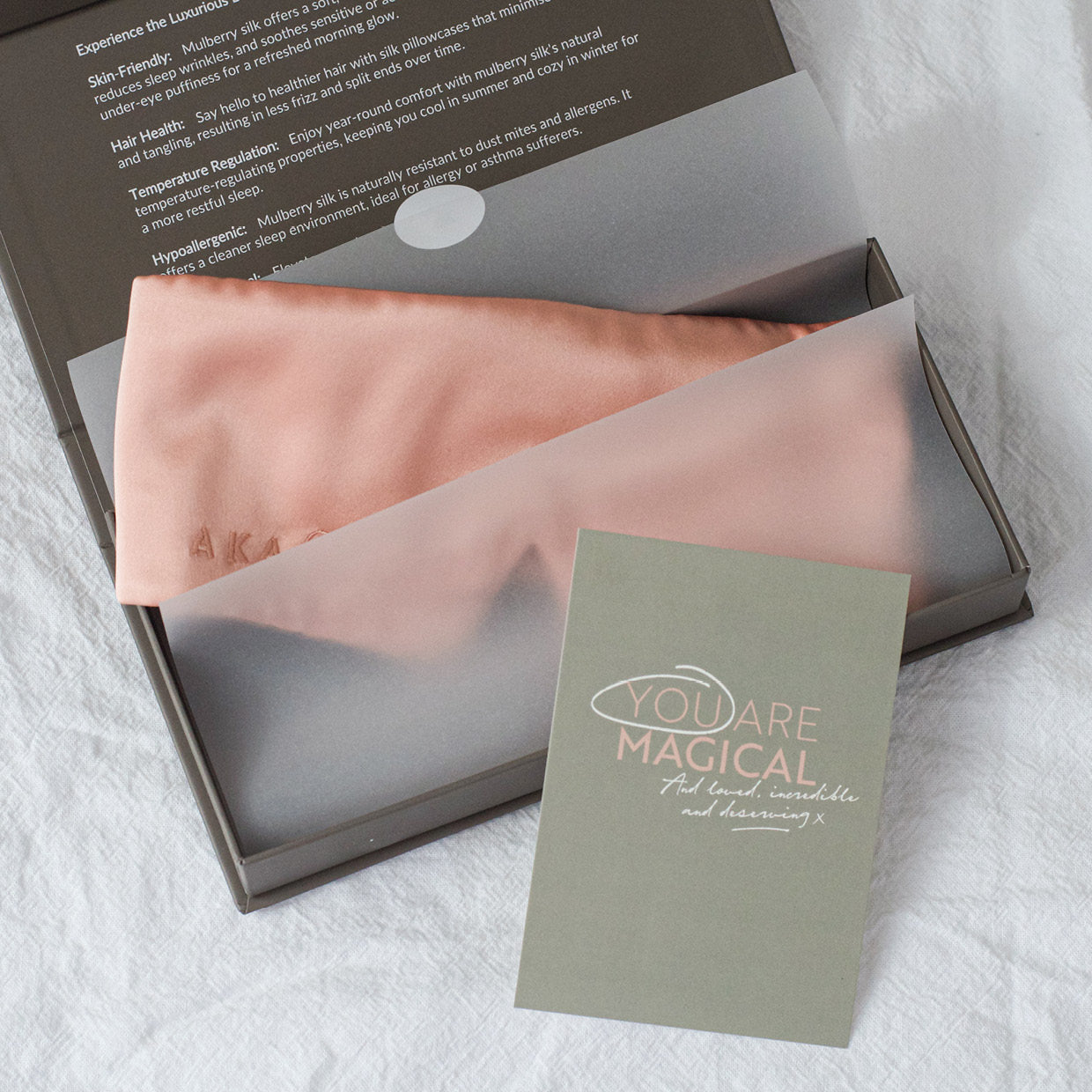 Blush Pink AKA Sleep Deep Sleep Eye Mask is lying in tissue paper in a gift ready charcoal gift box. There is a A5 light grey green card  propped in front of the box with the words "you are magical" written in all caps in light pink. In white script the words "And loved, incredible and deserving x" are underneath. The word YOU is circles in white to emphasize it.